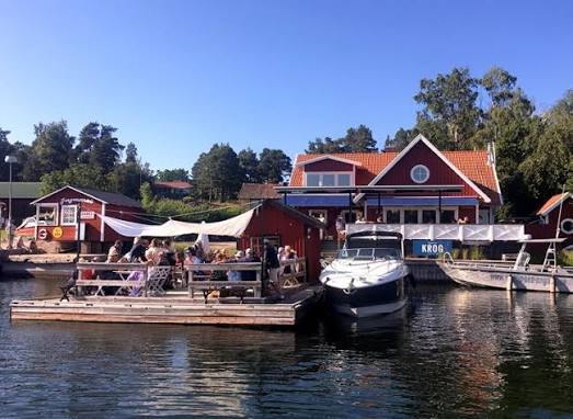 Ingmarsö Krog's red wooden restaurant with its KROG sign, a floating terrace full of guests and a motorboat at the jetty on Ingmarsö, Stockholm archipelago