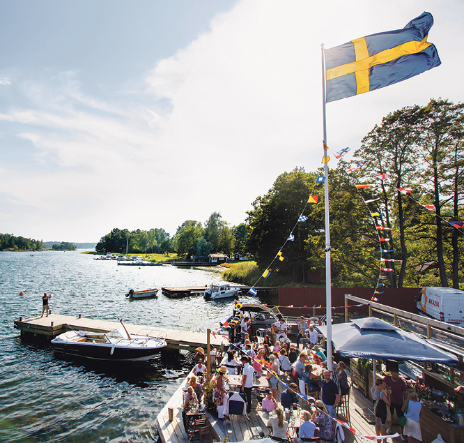 Busy jetty terrace under a Swedish flag and bunting at Motorverkstan Bistro & Bar on Djurö, with boats moored alongside, in the Stockholm archipelago
