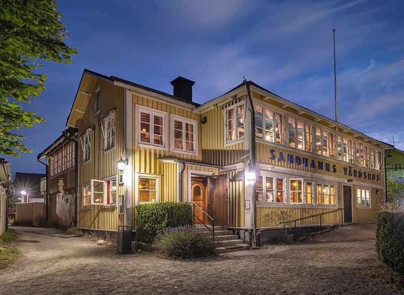Lit windows of the yellow two-storey Sandhamns Värdshus with its blue name sign at dusk, on Sandhamn in the Stockholm archipelago