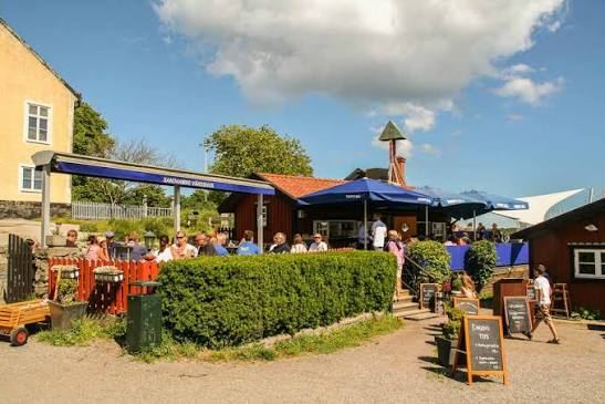 Guests at the outdoor terrace under the Sandhamns Värdshus awning, with blue parasols and chalkboard menus, on a sunny day on Sandhamn in the Stockholm archipelago