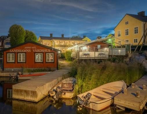 Red waterside building with the Sandhamns Värdshus Anno 1672 sign and small motorboats at a wooden jetty at dusk, on Sandhamn by boat in the Stockholm archipelago