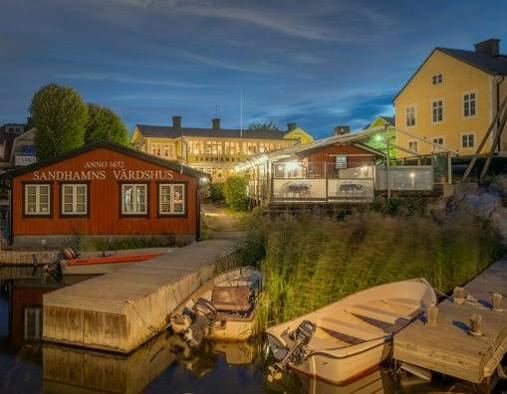 Red waterside building with the Sandhamns Värdshus Anno 1672 sign and small motorboats at a wooden jetty at dusk, on Sandhamn by boat in the Stockholm archipelago
