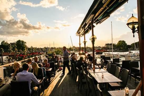 Guests dining on a sunlit wooden terrace with lanterns, looking out over the harbour and sailing masts at Seglarhotellet on Sandhamn, Stockholm archipelago