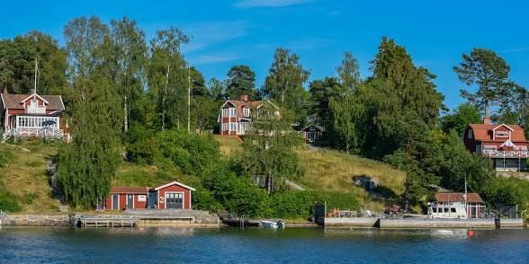 Red and white houses on a wooded hillside above the water on Blidö, seen from a boat in the Stockholm archipelago