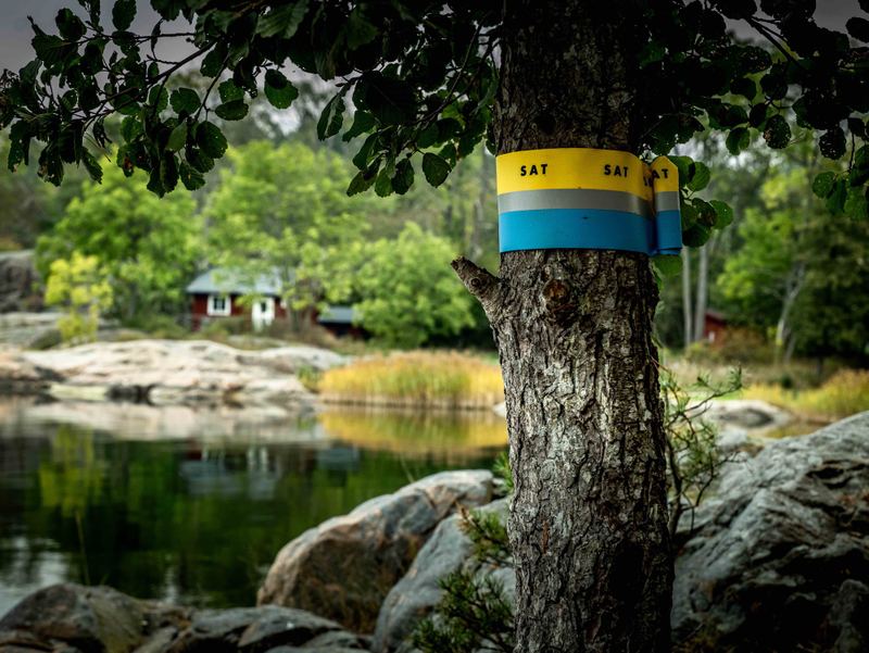 Yellow and blue trail marker on a tree beside a calm, rocky bay with a red cottage on Grinda