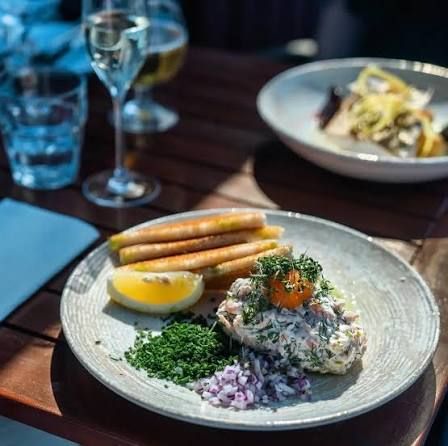 A plate of creamy salad topped with orange roe and dill, with rolled crispbread, lemon, chopped chives and red onion, and a glass of white wine, at Rökeriet on Fjäderholmarna near Stockholm