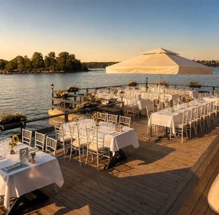 Tables with white tablecloths, white chairs and sunflowers on a wooden deck by the water at sunset, at Rökeriet on Fjäderholmarna in the Stockholm archipelago
