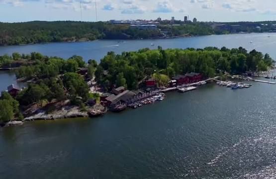 Aerial view of Fjäderholmarna with red buildings, jetties and moored boats along the shore and the Stockholm skyline beyond, where Rökeriet is reached by boat