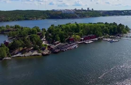 Aerial view of Fjäderholmarna with red buildings, jetties and moored boats along the shore and the Stockholm skyline beyond, where Rökeriet is reached by boat