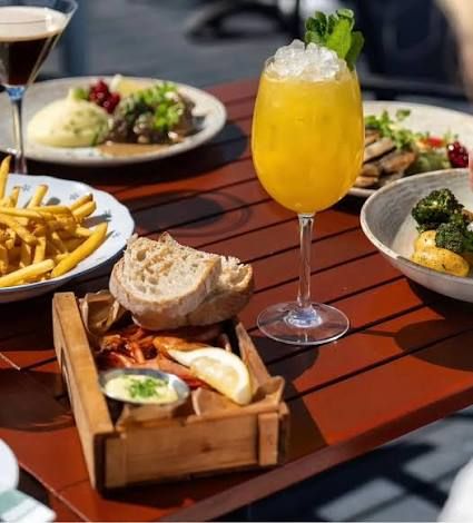 An orange cocktail with crushed ice and mint, bread, fried bites with lemon and a dip in a wooden tray, fries and other dishes on a table at Rökeriet on Fjäderholmarna near Stockholm