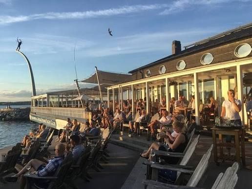 Guests in the evening sun on the terrace at Restaurang J in Nacka Strand, Stockholm, with the glass veranda and sculpture by the water