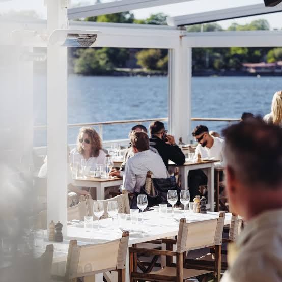 Diners at white tables on the veranda at Restaurang J, looking out over the water to wooded shores, a short trip by boat from central Stockholm