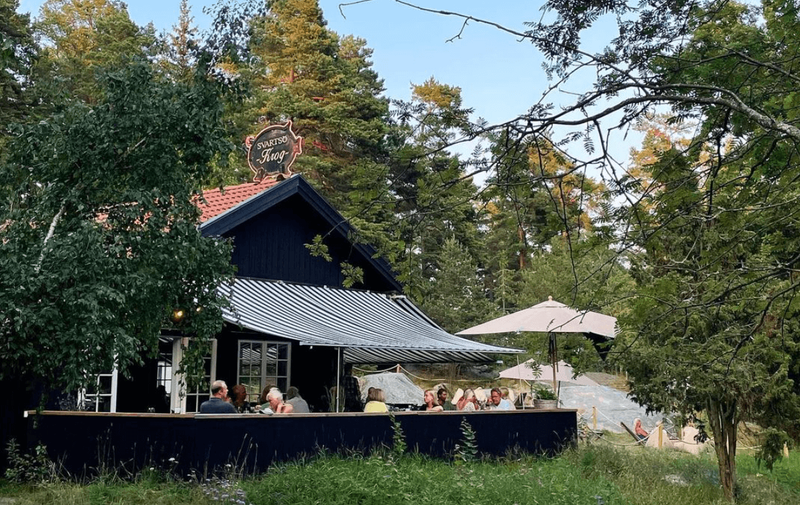 Black wooden house with a striped awning and outdoor dining among trees on Svartsö