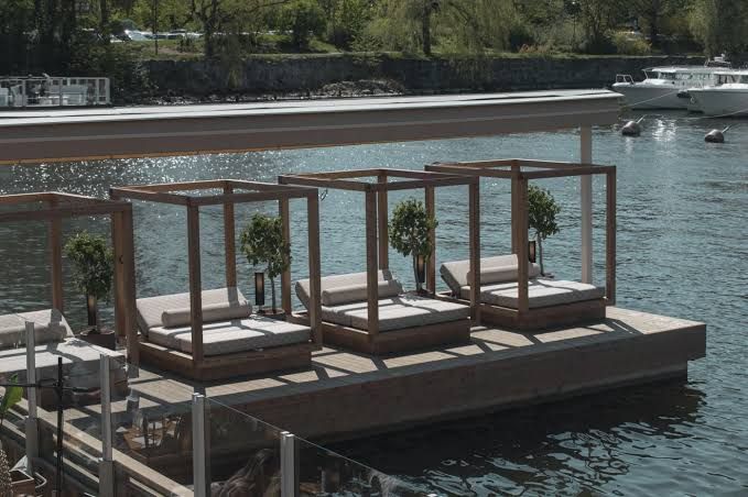 Wooden cabanas with cushioned daybeds on a deck over the water, with motorboats moored across the bay, at Strandbryggan in Stockholm