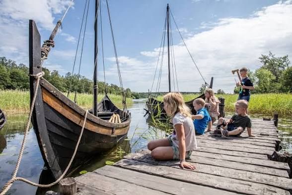 Children sitting on a wooden jetty beside replica Viking ships at Birka