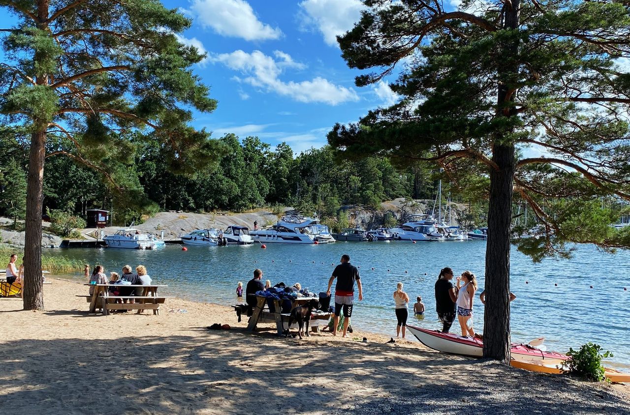 Sandy beach on Finnhamn with people at a picnic table and boats moored across the bay