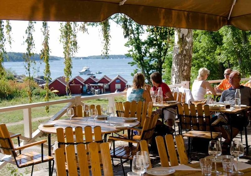 Guests dining on the veranda at Grinda Wärdshus, looking out over red boathouses and the water