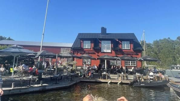 Guests under parasols on the waterfront terrace in front of the red Motorverkstan building on Djurö, seen by boat in the Stockholm archipelago