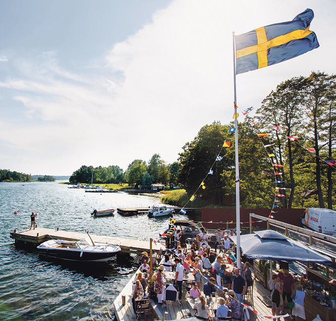 Guests on the flag-decked jetty terrace at Motorverkstan Bistro & Bar on Djurö, with motorboats at the jetties, in the Stockholm archipelago
