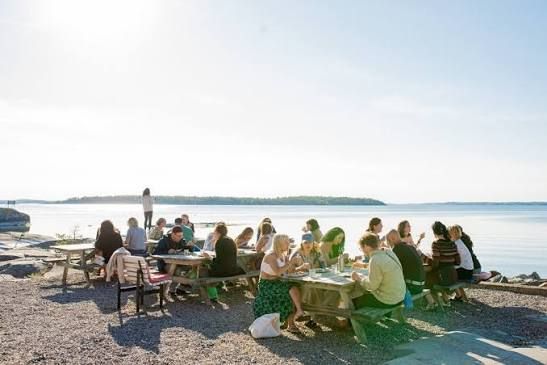 Guests at wooden picnic tables on the gravel shore at Grundet on Rögrund, looking out over sunlit water in the Stockholm archipelago