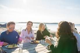 Friends sharing plates of food at an outdoor wooden table by the water in the sun at Grundet in the Stockholm archipelago