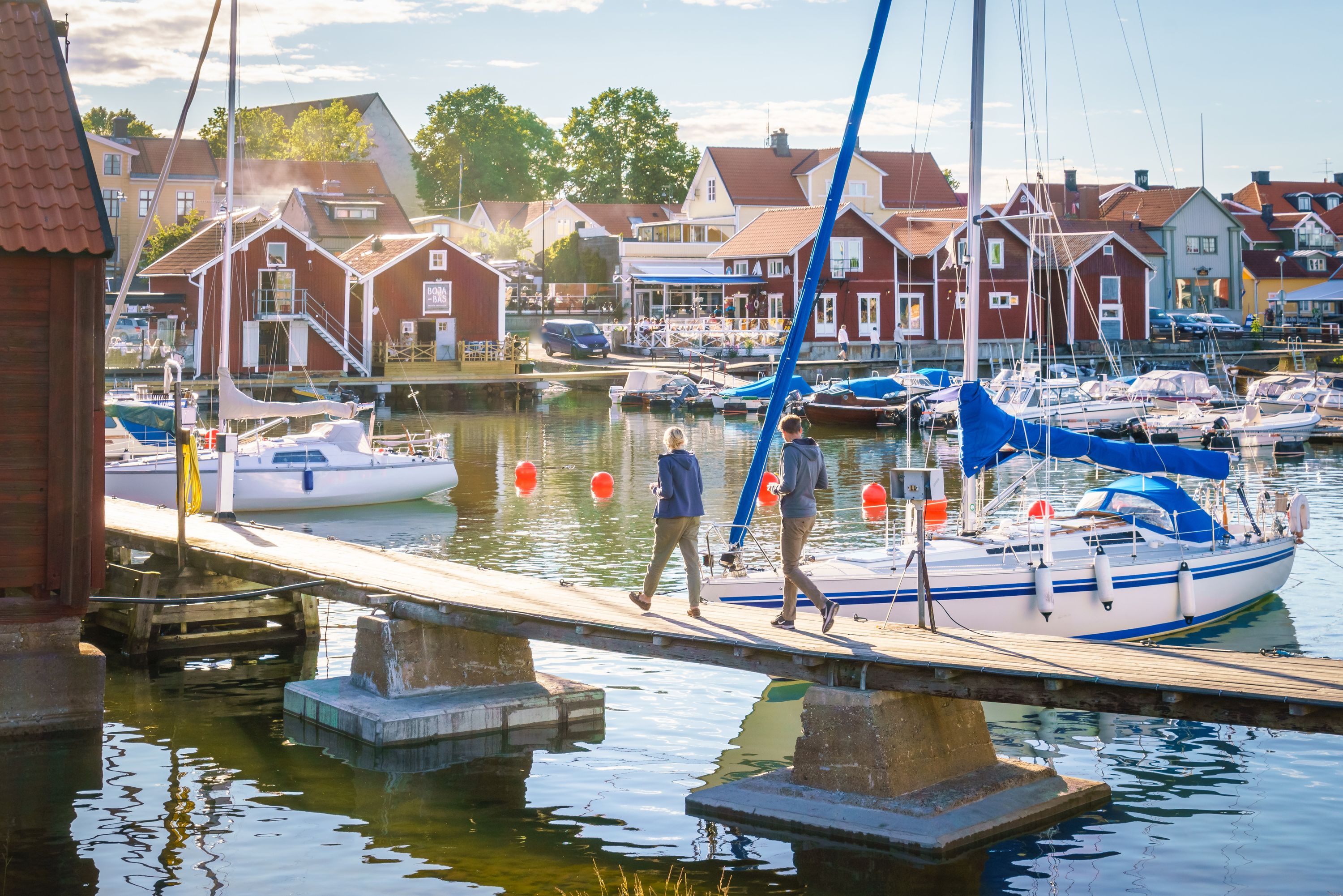 People walking along a jetty past sailboats in a Möja harbour lined with red wooden houses