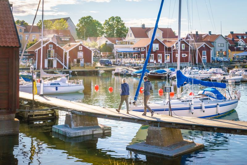 People walking along a jetty past sailboats in a Möja harbour lined with red wooden houses