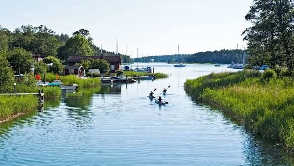 Kayakers paddling along a reed-lined channel past houses and moored boats on Utö